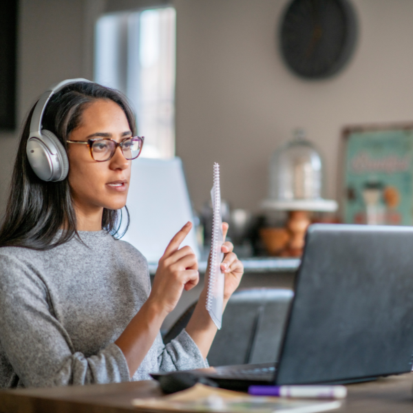 A woman wearing over the ear headphones is holding a notepad and pointing to it while looking at a laptop screen