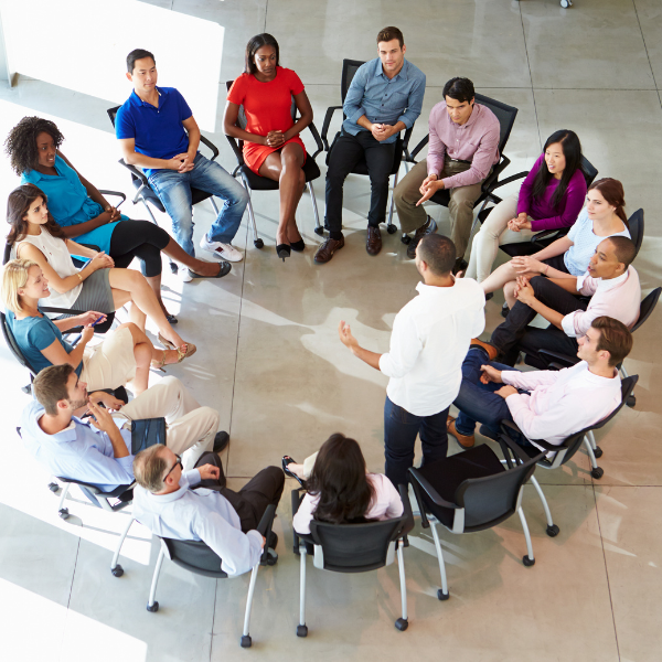 A group of 15 adults sit in a circle listening to a man in a white shirt standing up