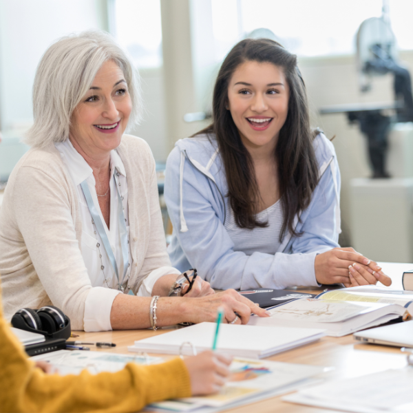 Two women are smiling while seated at a table of people examining papers and writing