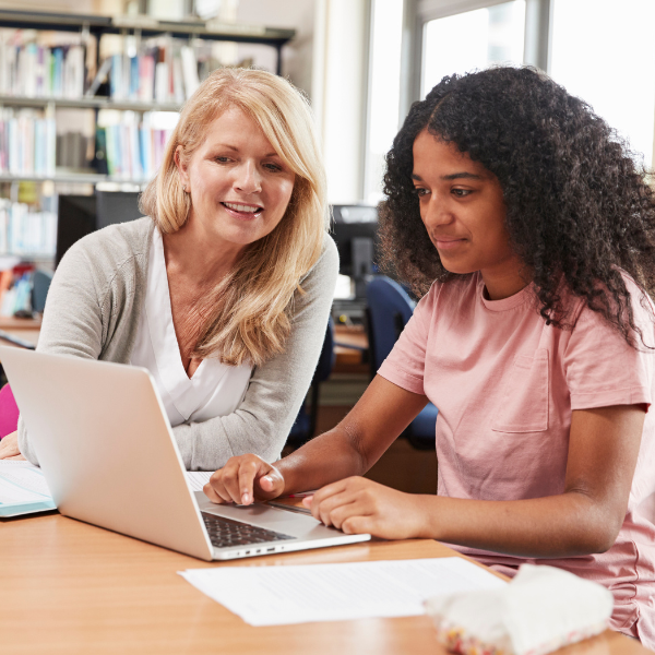 A female teacher sits next to a teenage student who is sitting in front of a laptop smiling