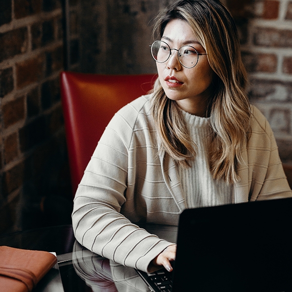 A woman wearing glasses and a tan seater sits at a laptop looking to the side