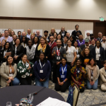 The School Teams AI Collaborative at the convening in Denver, Colorado. It is a large group photo taken in a conference room. Approximately 70-80 diverse professionals are gathered together, arranged in multiple rows, with some people standing and others kneeling in the front.