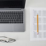 A workspace setup showing a laptop with a black keyboard, a pair of glasses, and a standardized test answer sheet or bubble sheet with a yellow pencil placed on top. The items are arranged on a white surface, creating a clean, academic or testing environment composition.