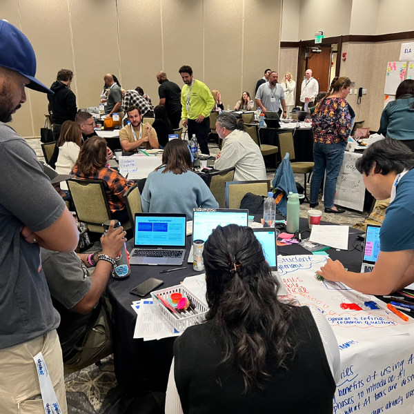 A conference room filled with School Teams AI Collaborative members working in groups. School leaders are seated around tables with laptops, posters, and markers, engaging in discussions around AI. The room has a professional setting with beige walls.