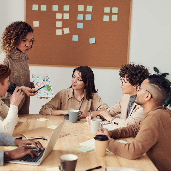 A group of researchers gathered around a wooden table building collaborative research partnerships. One person is standing and presenting data on a tablet, while others sit attentively with coffee cups, notebooks, and a laptop. A corkboard with colorful sticky notes is visible in the background, suggesting brainstorming or planning activities.